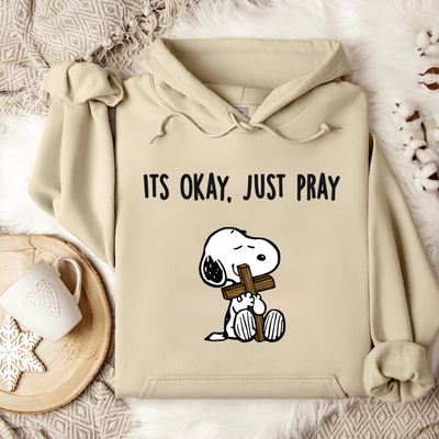 Beige hoodie with cartoon character holding a cross and text 'ITS OKAY, JUST PRAY' on a soft surface.